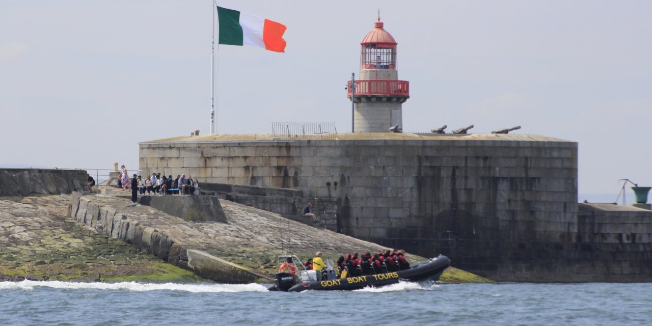 Full boat of people sailing in front of a light house 